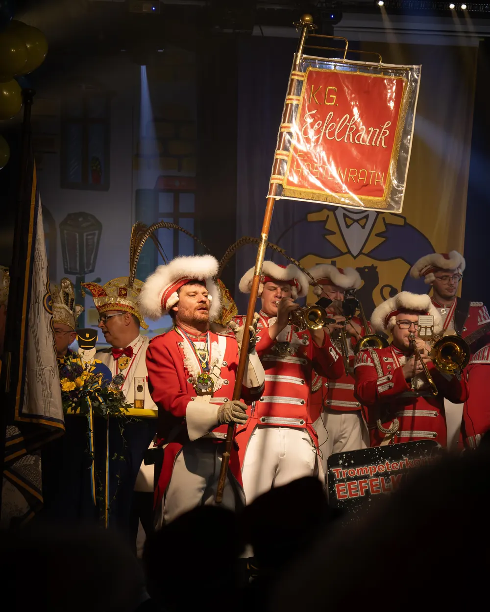 Ein Musiker in Uniform trägt die Fahne der KG Eefelkank, dahinter das Trompeterkorps